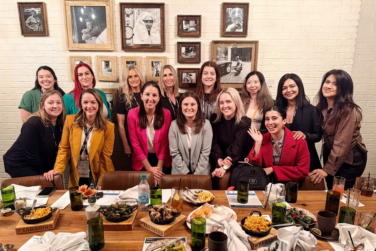 Own the Mic Lunch A group of women pose for a photo at the “Own the Mic” media training lunch, standing behind a long table with shared plates and drinks in a restaurant.